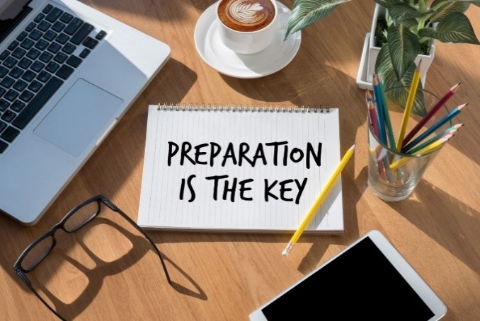 A notebook with "Preparation is the key" written it sits on a desk, with a laptop, coffee, pencils, a plant and glasses surrounding it.