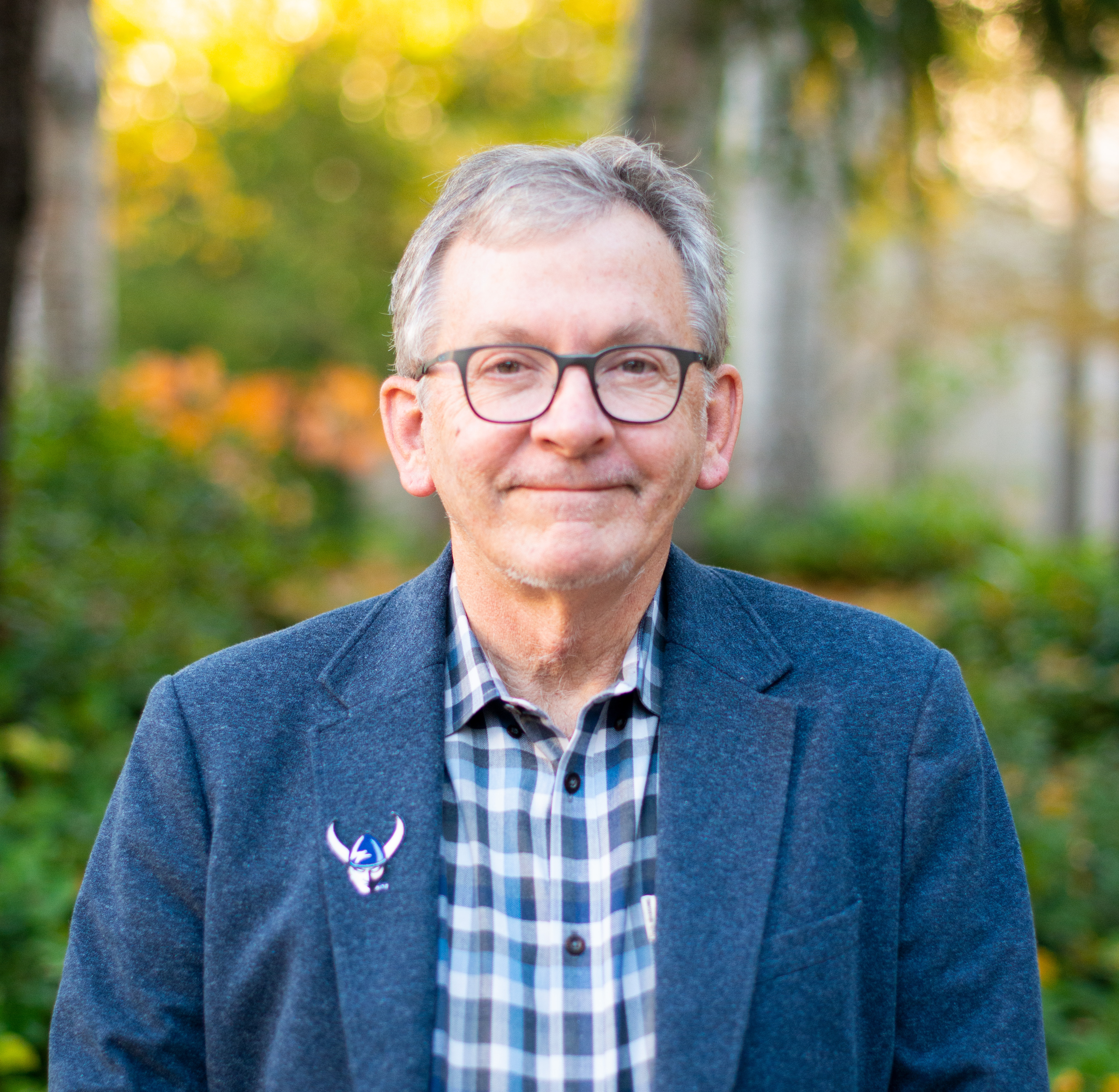 Dean Scott T. young in a blue check skirt and sports coat with a viking pin, standing on an outdoor campus walkway