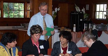 A man holding a green piece of paper standing behind a group of women sitting at a table