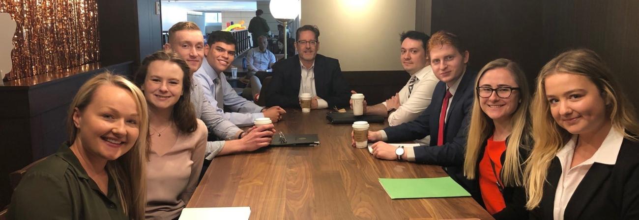 Financial Management Association students sit at a table during a meeting holding paper coffee cups wearing business attire.