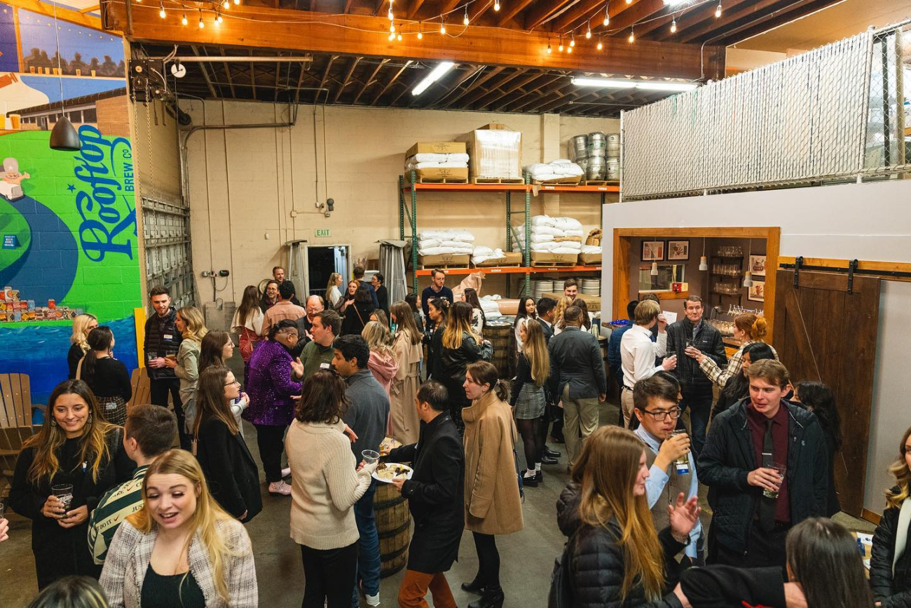 A group of people gathered in a warehouse-like space for the Holiday Mixer. The people are standing and mingling, some are holding drinks. There are stacks of boxes and shelves in the background.