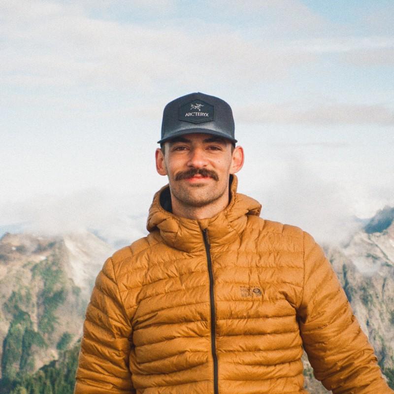 Photo of Clay Ashworth in mustard yellow zip-up jacket and a dark hat, in front mountain range. 